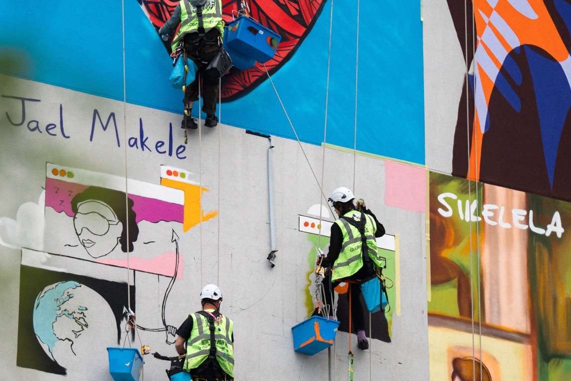 Two painters in harnesses and hard hats working on a large mural suspended from ropes