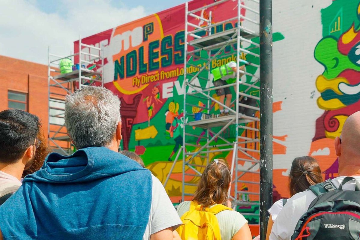 A group of people standing in the street watching a large mural being painted from scaffolding