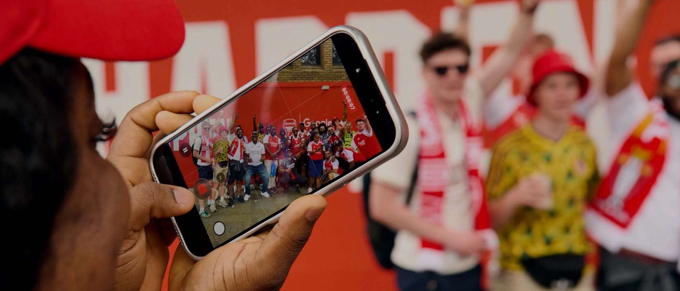 A phone held up photographing a crowd of people standing in front of a painted wall