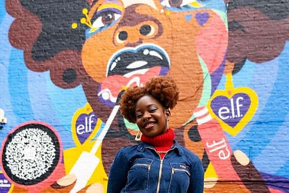 A person standing smiling in front of a brightly coloured hand-painted wall